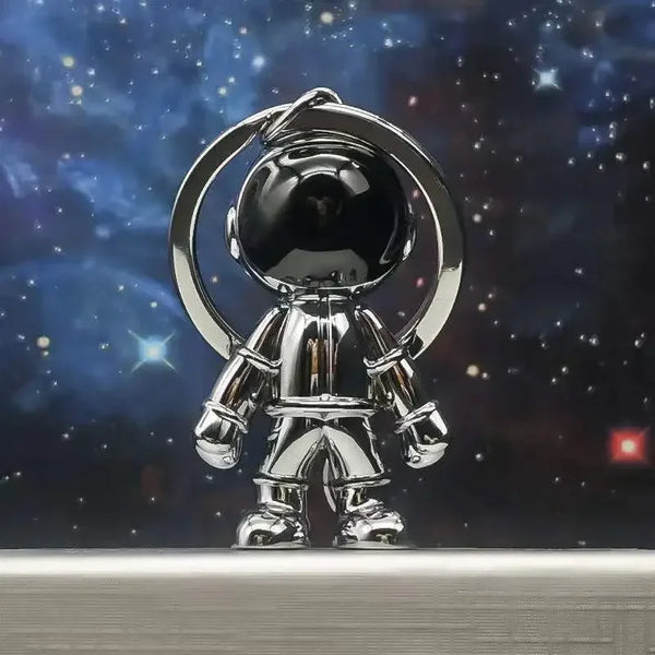 Colgante de llavero plateado brillante con casco y traje espacial contra fondo estrellado oscuro.