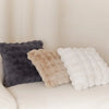 Three fluffy decorative pillows in shades of gray, beige, and white resting on a light-colored sofa.