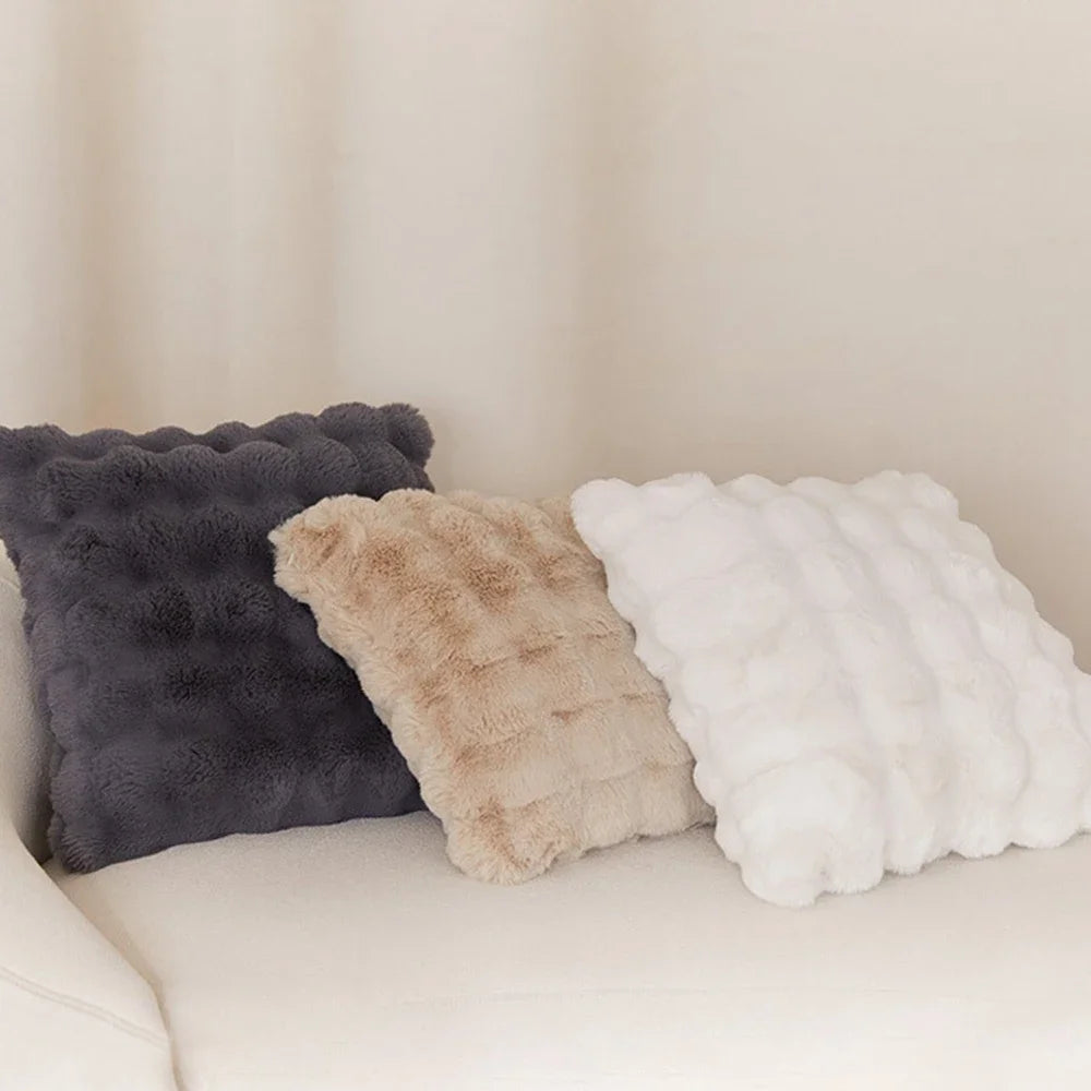 Three fluffy decorative pillows in shades of gray, beige, and white resting on a light-colored sofa.