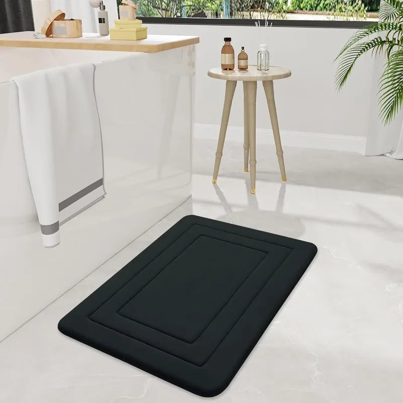 A black absorbent non-slip bath mat is placed on a white floor next to a counter and a small wooden table.