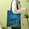 A person holds a blue Van Gogh series canvas tote bag featuring a starry night landscape on water.