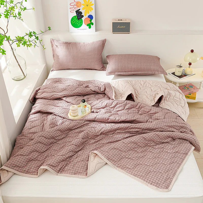 A light pink quilted blanket is displayed on a bed with matching pillows and a small plant in the background.
