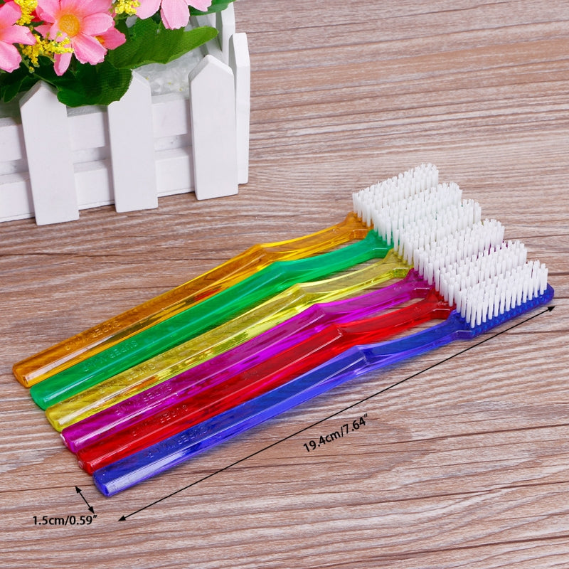 A set of six colorful hard bristled toothbrushes laid out on a wooden surface next to a white picket fence and pink flowers.