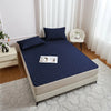 A navy blue waterproof mattress protector with elastic straps is displayed on a bed, next to a white chair and light-colored 