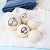 A set of six white wool dryer balls with embroidered sheep faces resting on a white fabric.