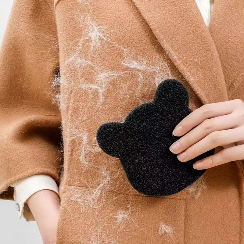 Une main tient une éponge retire-poils noire contre un tissu de blazer beige avec des fibres de cheveux blancs visibles.