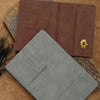 A brown leather passport holder and a gray leather cardholder are displayed on a tan fabric background.