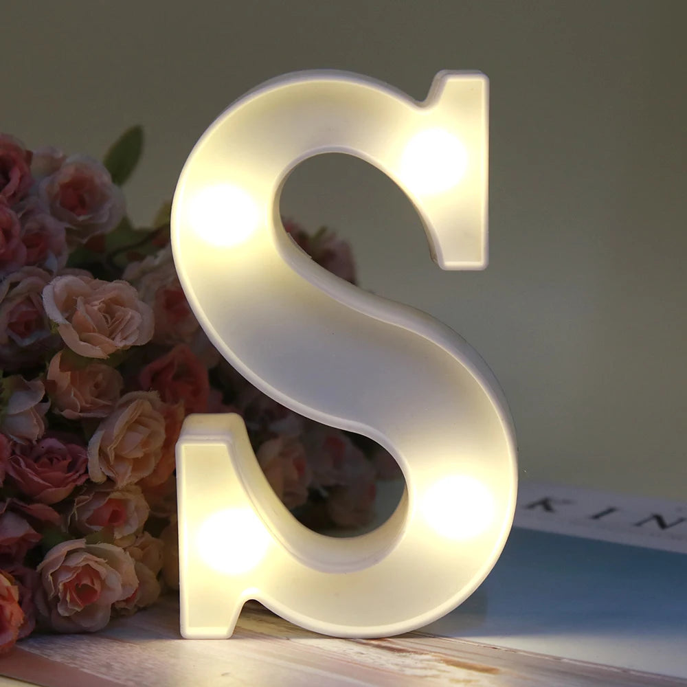 Lumière de nuit alphabétique en LED blanche en forme de lettre S, posée sur une surface en bois à côté de roses roses.
