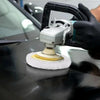 A hand wearing a glove is using a polishing tool with a wool disc on a dark car surface.
