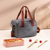 A gray compact thermal lunch tote bag with brown straps rests on a white surface next to cutlery.