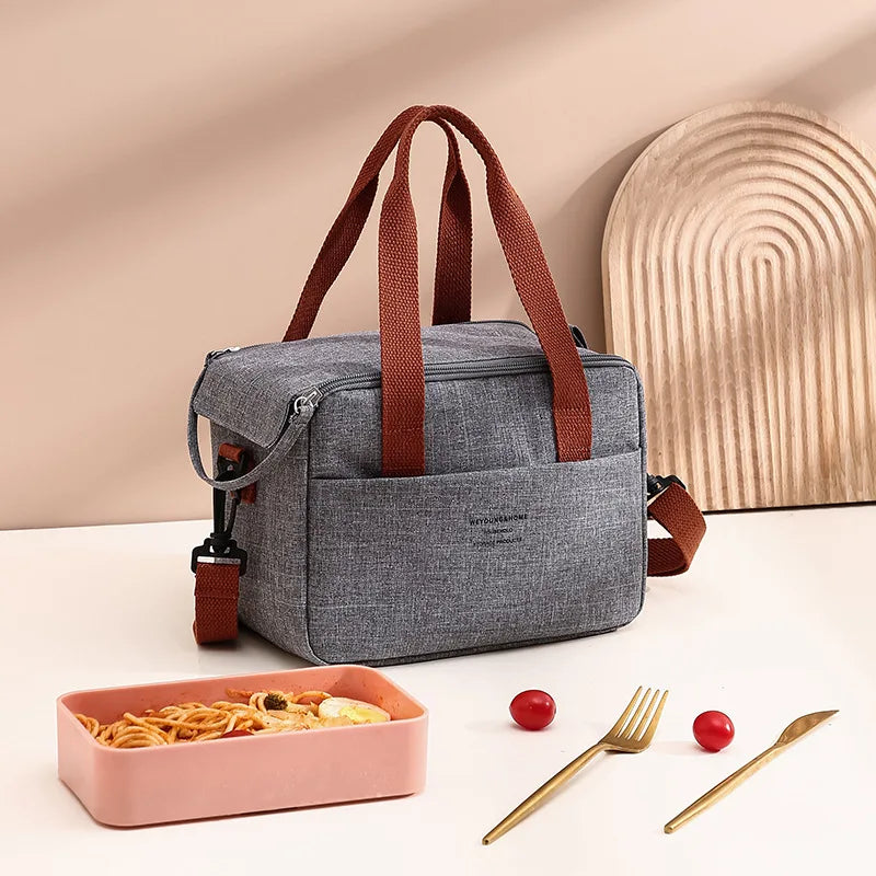 A gray compact thermal lunch tote bag with brown straps rests on a white surface next to cutlery.