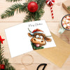 A Christmas card featuring an adorable Highland cow illustration on a white card with a kraft paper envelope and festive gree