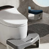 A white comfort squatting stool with a gray footrest is placed on a tiled floor next to a window.