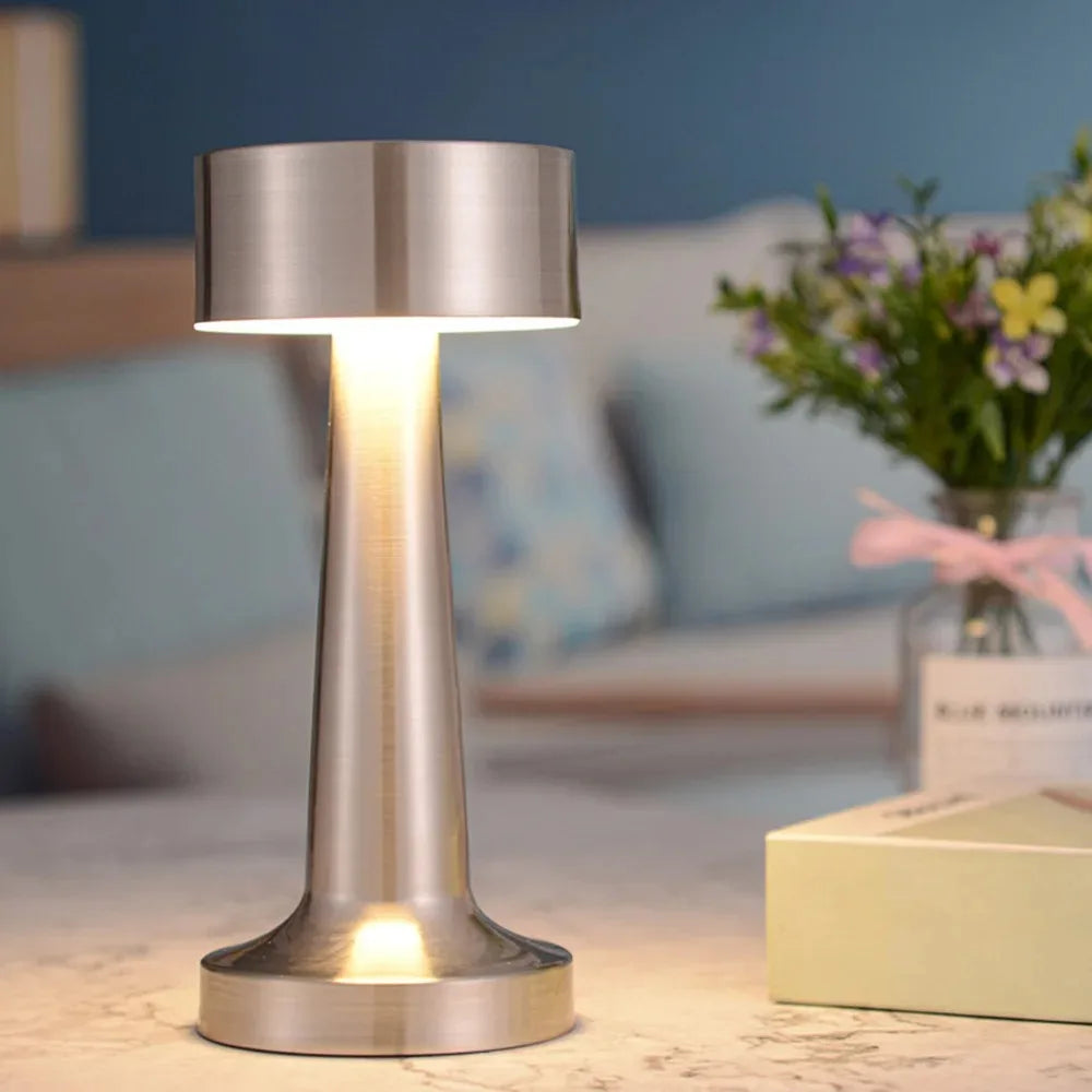 A modern silver metal LED table lamp with a cylindrical shade illuminates a surface in a softly lit indoor setting.