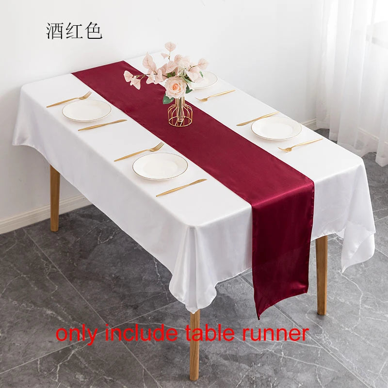 A white tablecloth set with a deep burgundy satin table runner, gold cutlery, and floral centerpiece.