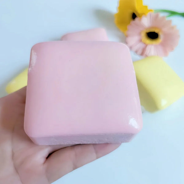 A hand holds a square pink squishy toy shaped like a mousse cake, with yellow flowers in the background.