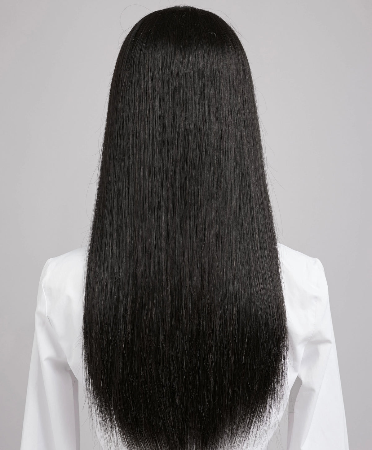 Una larga peluca de cabello negro de seda lisa, mostrada desde atrás contra un fondo gris claro.