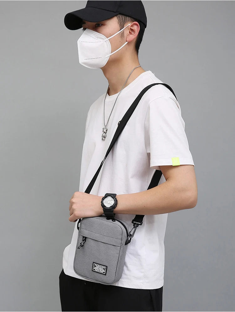 A person wearing a black cap and face mask holds a gray mini crossbody phone bag with an adjustable strap.