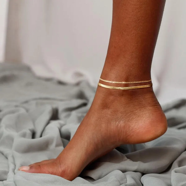 A close-up of a person's ankle showing a layered gold herringbone anklet set on the skin.