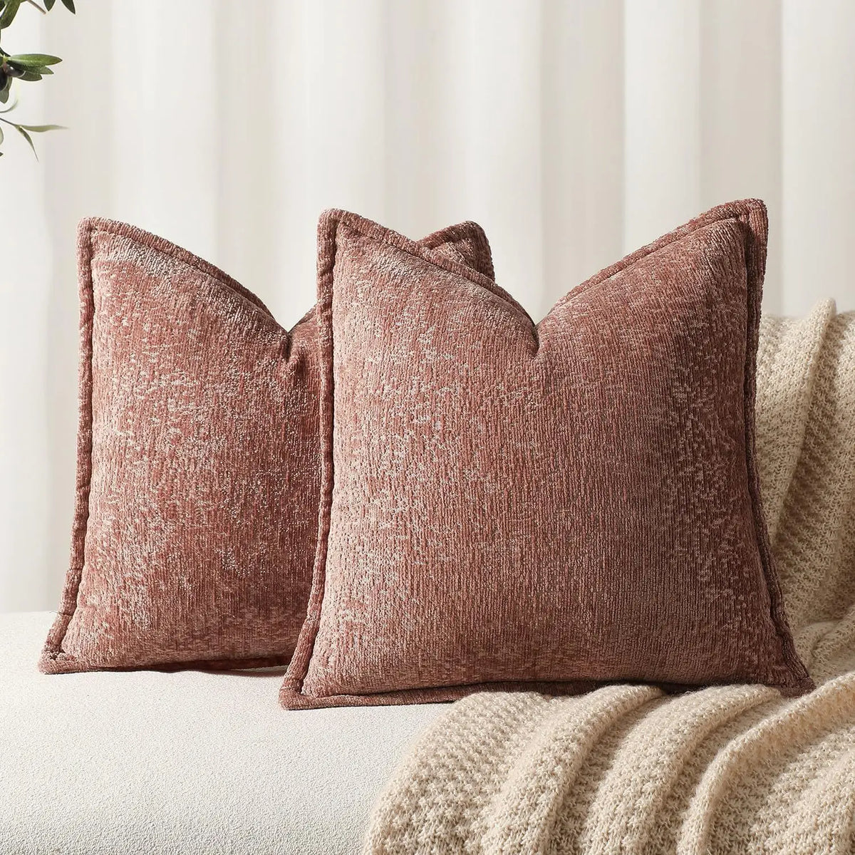 A square, textured, dusty rose chenille decorative cushion cover resting on a beige knitted throw blanket.