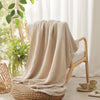 A beige soft muslin cotton blanket draped over a wooden chair and resting on a woven seat.