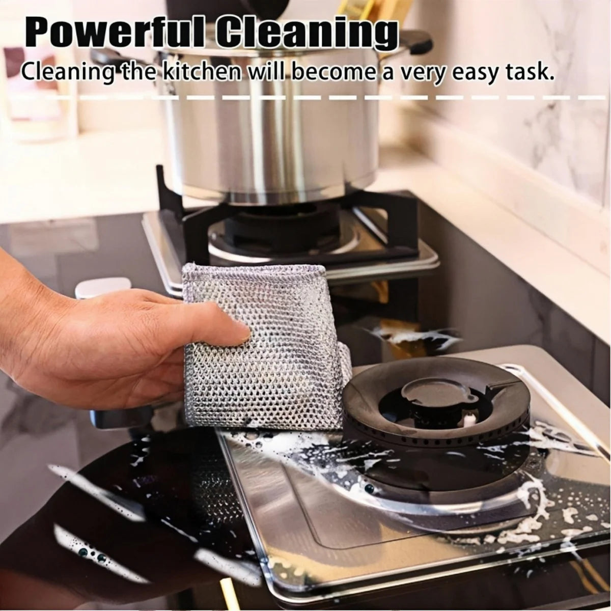A hand is using a gray scouring cloth to clean a black non-stick pan on a stovetop.