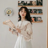 A young woman wearing a cream-colored vintage lace long sleeve blouse and white skirt, holding a newspaper.