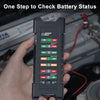 A hand holds a black car battery tester device displaying various LED indicators for battery status.