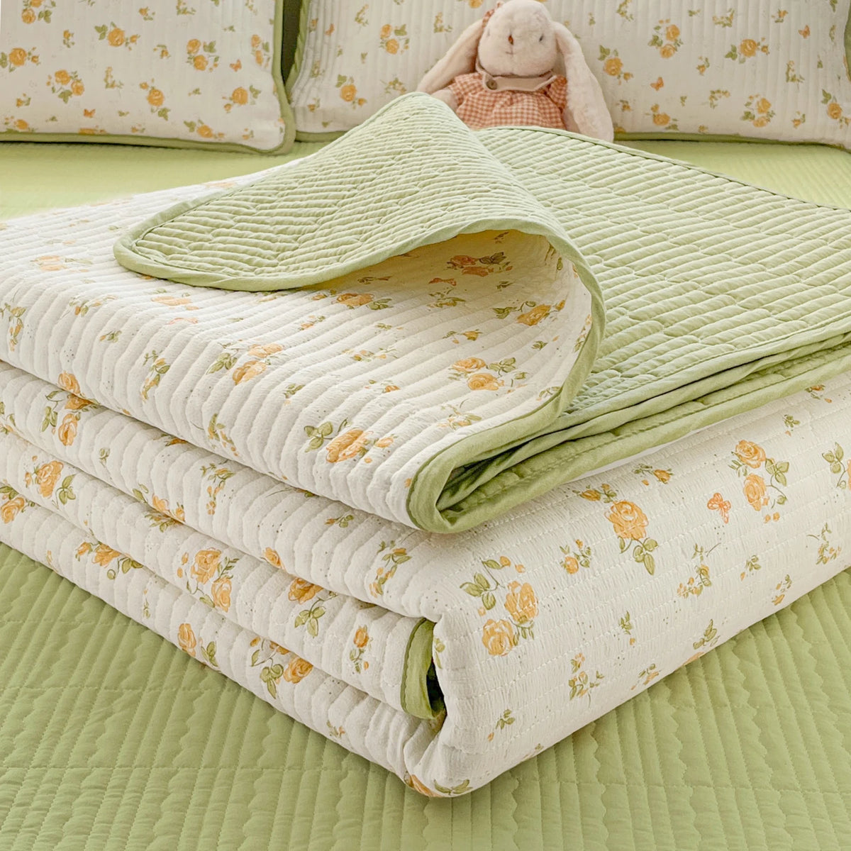 A stack of floral quilted bedspreads in light green and cream colors with a small stuffed animal on top.