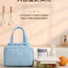 A light blue insulated quilted lunch tote bag is shown next to a clear food container on a wooden table.