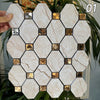 A hand holds a square mosaic tile featuring beige marble pattern and gold accents, set against green foliage.