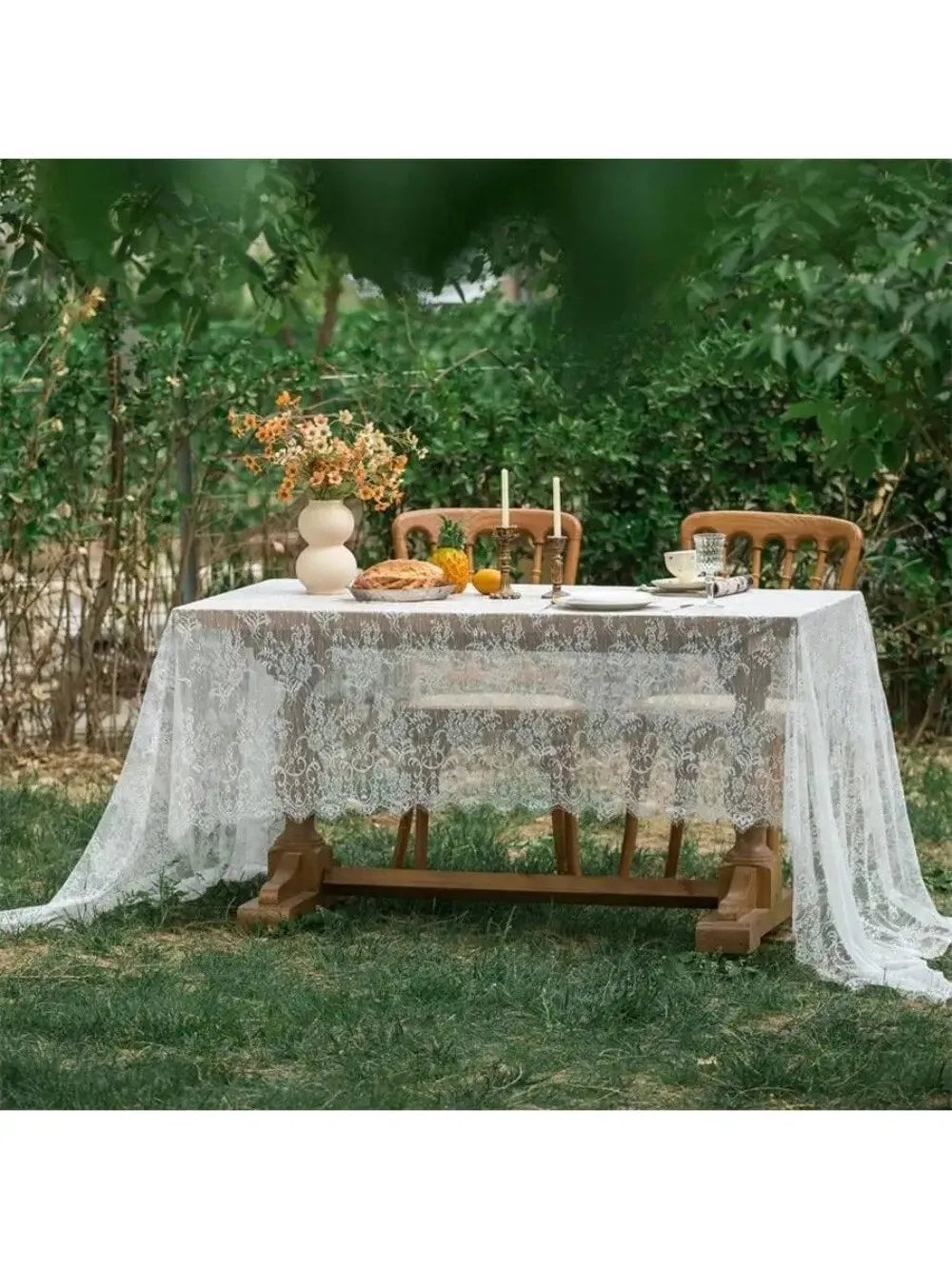 Elegant white lace tablecloth set up outdoors on a wooden table with floral decorations.