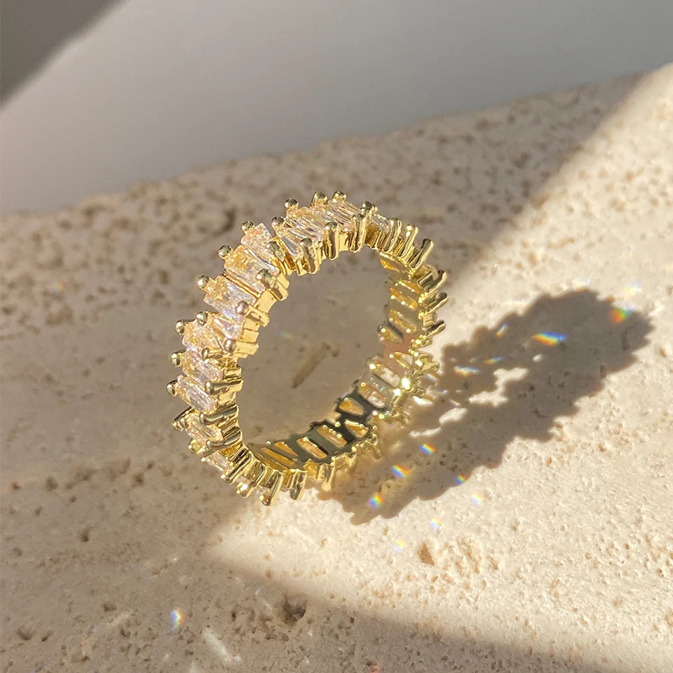 A close-up of a gold ring featuring a row of baguette cubic zirconia stones, resting on a light surface.
