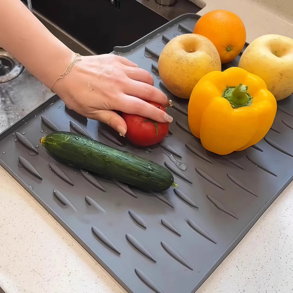 A hand places various fresh produce like apples, a bell pepper, and a cucumber onto a silicone sink drain mat.