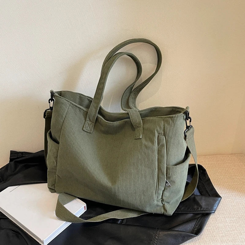 A olive green corduroy tote bag with a leather strap resting on a black surface next to a white book.