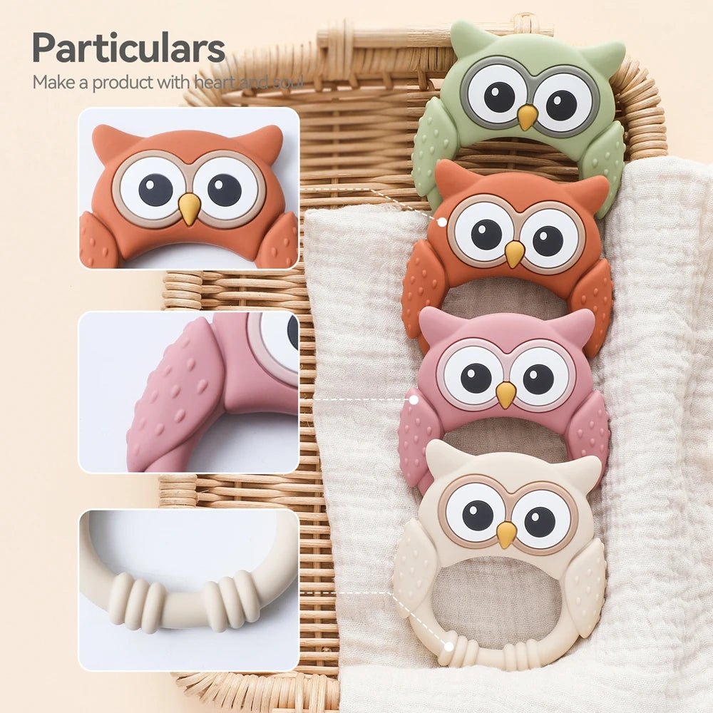 A set of three owl-shaped silicone teethers and rattles in orange, pink, and beige are displayed on a woven tray.