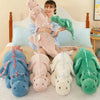 A woman is posing with various soft stuffed dragon plush toys in shades of pink, blue, and green on a bed.