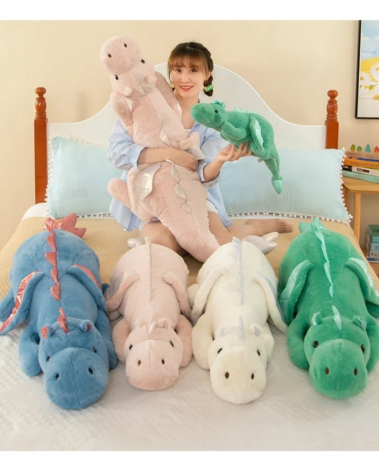 A woman is posing with various soft stuffed dragon plush toys in shades of pink, blue, and green on a bed.