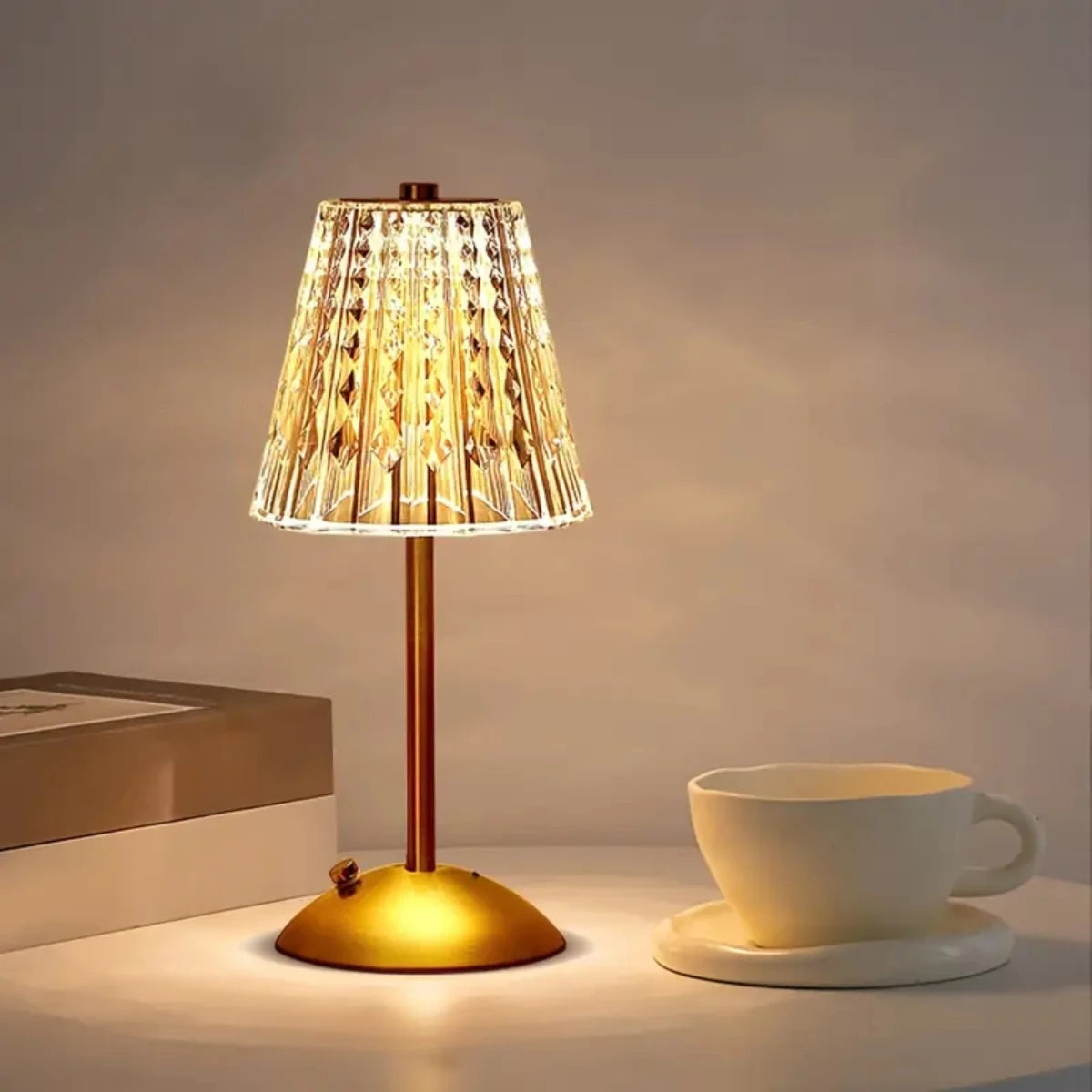 A gold-toned table lamp with a textured glass shade illuminates a white cup on a light surface.