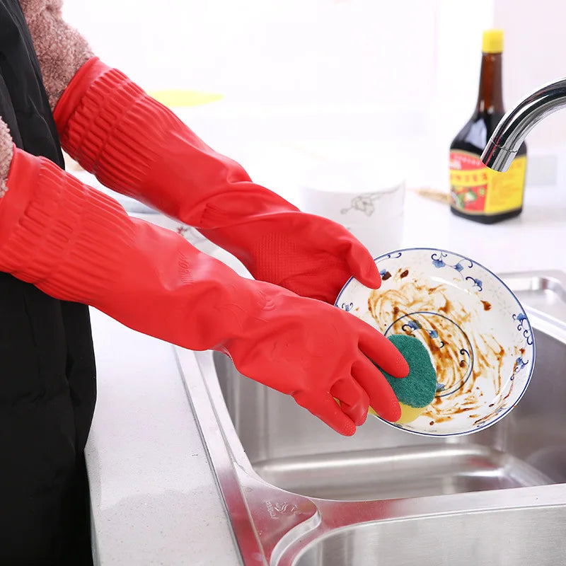 A pair of red silicone scrubbing gloves are shown dipping into a stainless steel sink for dishwashing.