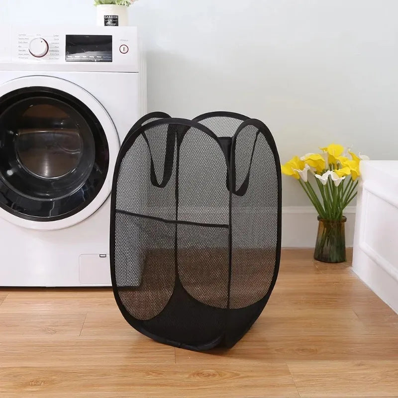 A black foldable laundry hamper with a mesh design sits on a wooden floor next to a white washing machine.