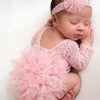 A newborn baby wearing a pink lace romper with ruffles and a matching headband, lying on a white surface.