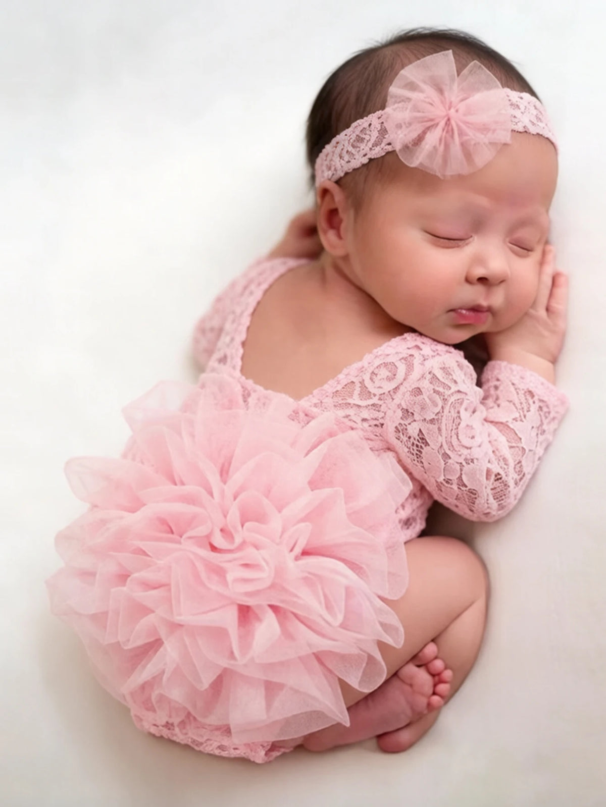 A newborn baby wearing a pink lace romper with ruffles and a matching headband, lying on a white surface.