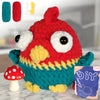 A red and green crocheted bird amigurumi toy with large white eyes sitting next to a red and yellow yarn kit.