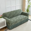 A moss green jacquard sofa cover with an elastic skirt displayed indoors against a white wall.