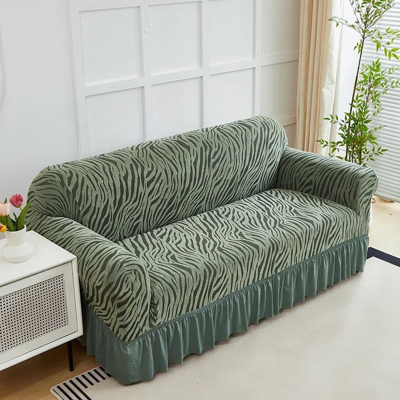 A moss green jacquard sofa cover with an elastic skirt displayed indoors against a white wall.