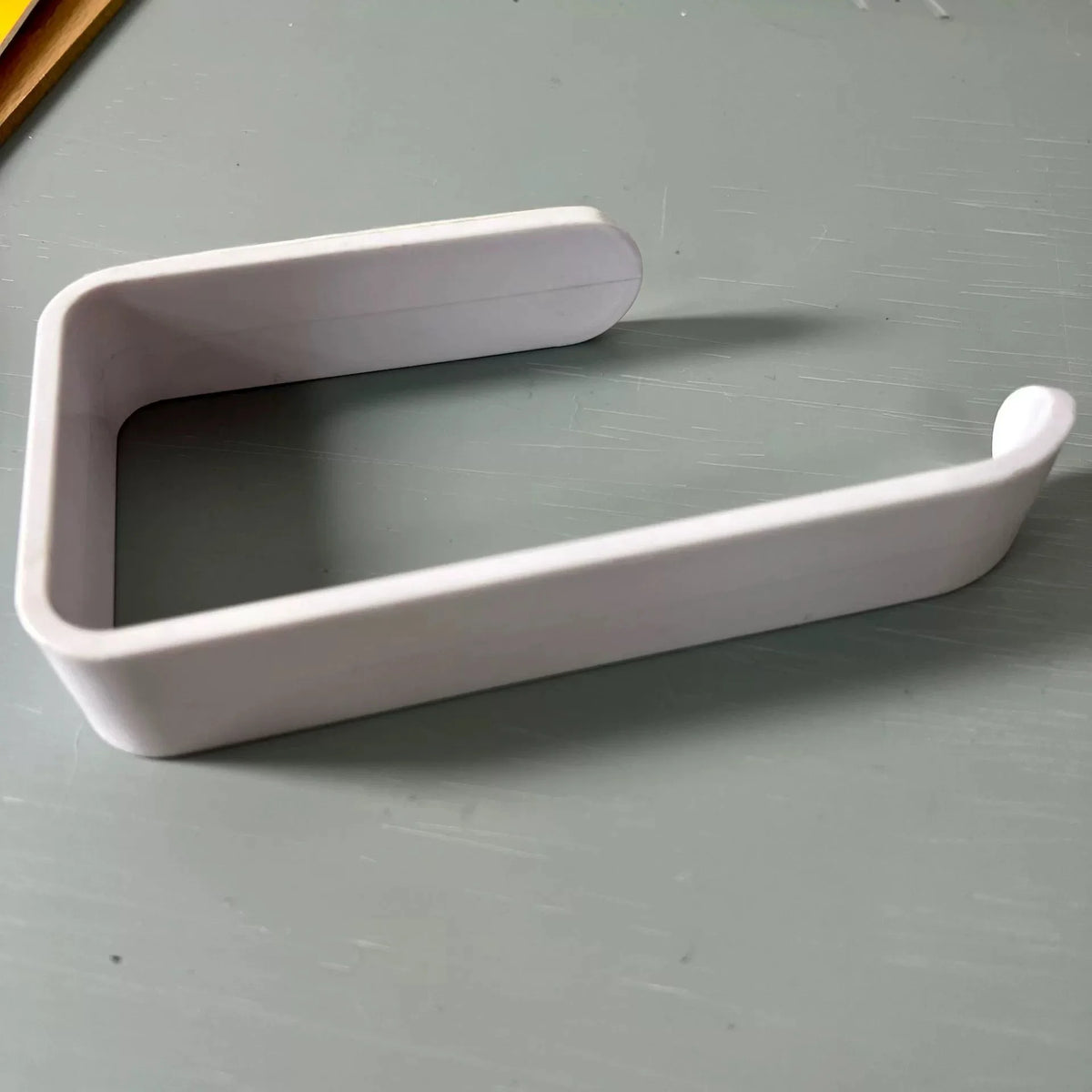 A white No-Drill Self-Adhesive Toilet Paper Holder with a curved design shown on a gray surface.
