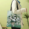 A person holds a light green canvas tote bag featuring a white floral painting, while wearing a cream blazer and blue shirt.