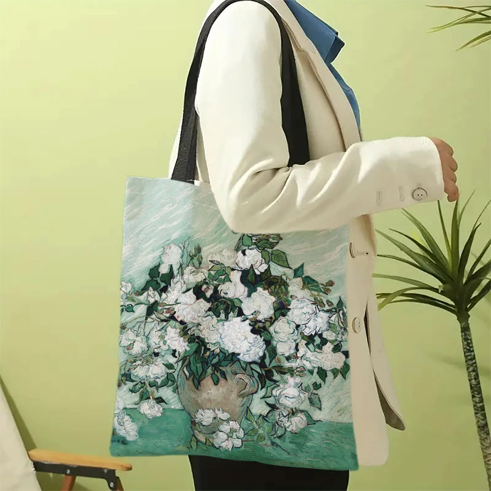 A person holds a light green canvas tote bag featuring a white floral painting, while wearing a cream blazer and blue shirt.