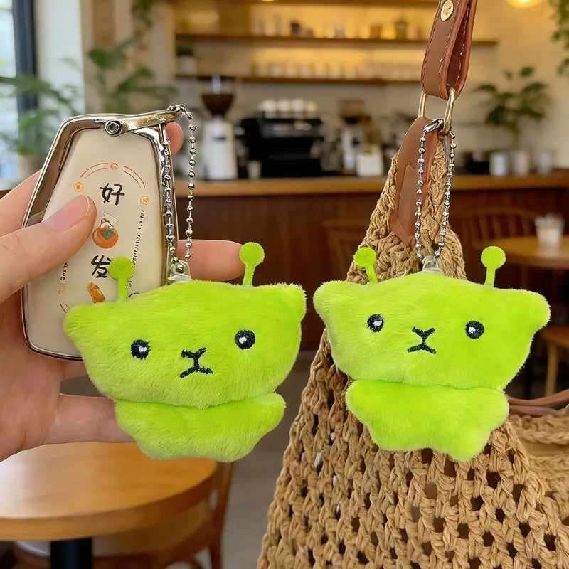 A bright lime green alien plush charm with small antennae is attached to a leather keychain and displayed next to a woven bag